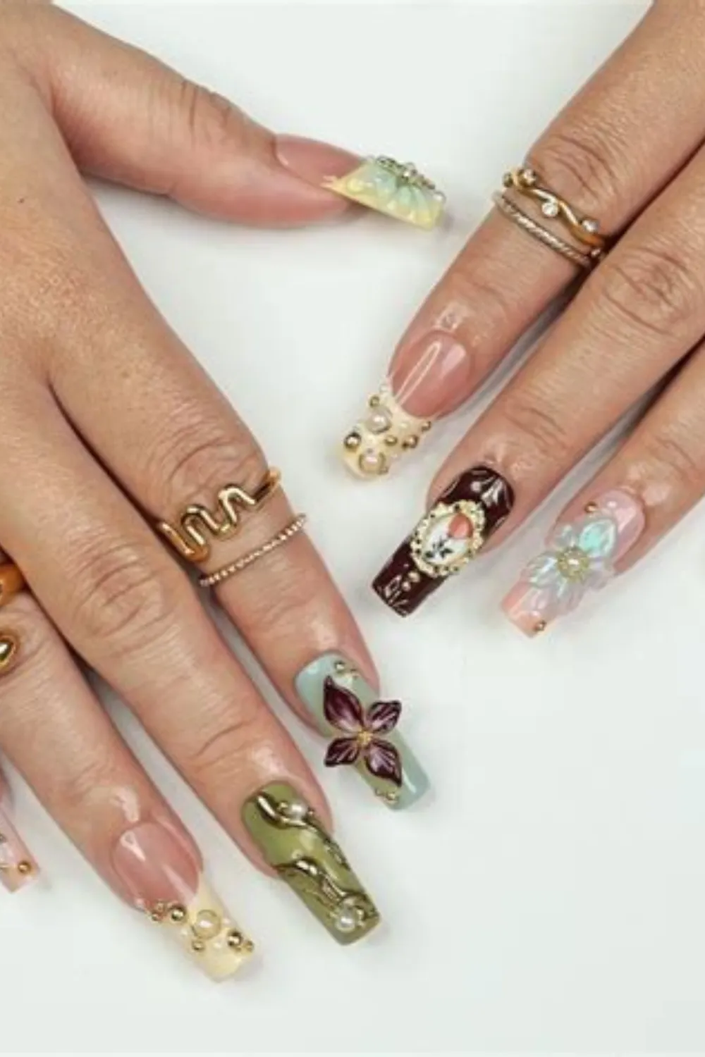 A close-up photograph of two hands displaying a vintage, antique-inspired maximalist manicure on long square nails. The design features a mix of French tips, olive green swirl art, a dark chocolate-brown accent nail framed with an antique oil painting frame, and 3D sculpted iridescent flower charms accompanied by gold studs and tiny pearls.