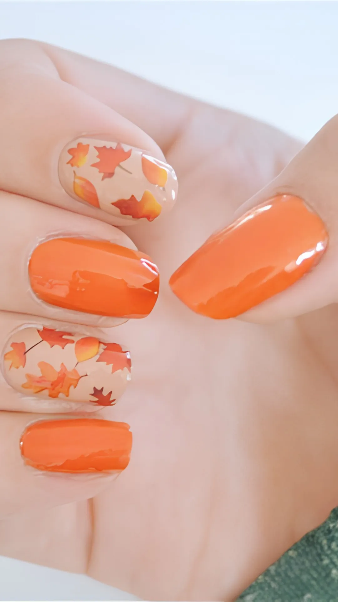 Vibrant Thanksgiving nail art showing a combination of solid, glossy orange nails and neutral nude nails decorated with drifting red and orange maple leaves.