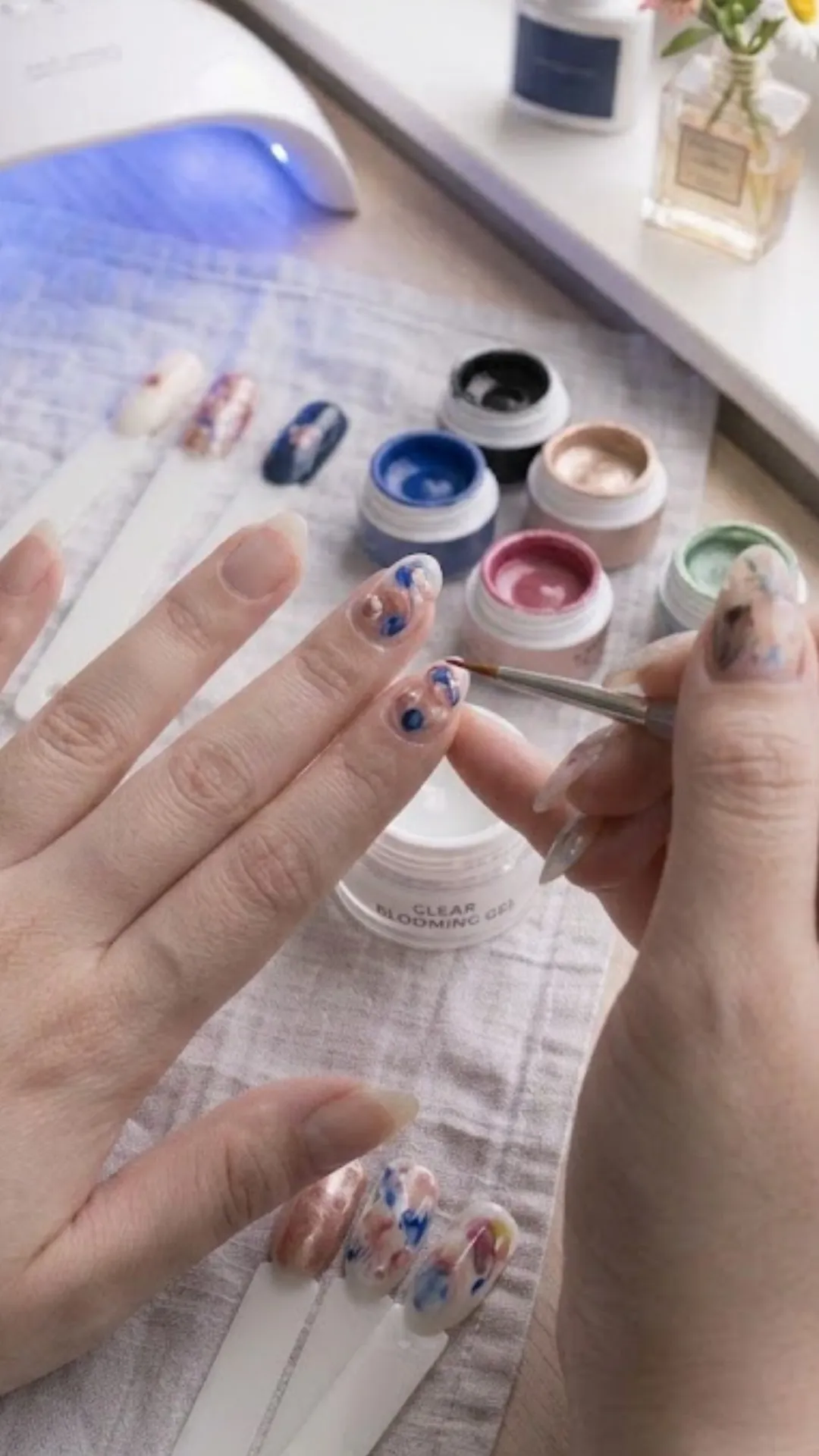 A close-up of the watercolor painting process on natural nails, showing a fine-tip brush blending blue and copper metallic gel polishes on a wet blooming gel layer to create an organic marble and floral pattern.