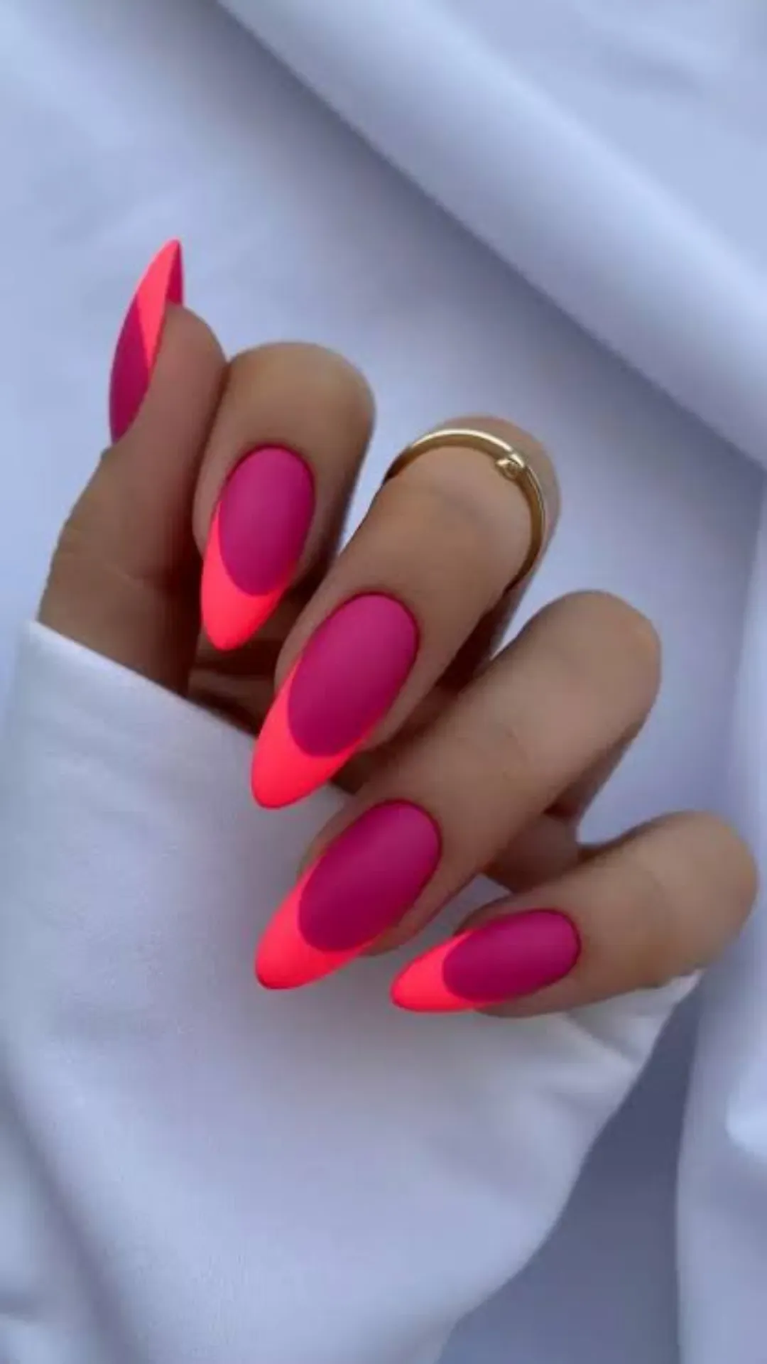 A close-up view focuses on a hand with vibrant, matte magenta almond-shaped nails, featuring a dynamic neon coral diagonal tip design. The manicure is clean and sharply defined, creating a modern, bold alternative to traditional everyday nails. The hand wears a single simple gold ring on the index finger and rests against a contrasting white fabric, emphasizing the bright, cheerful colors and velvety matte finish. This striking gel nail art provides an energetic yet sophisticated look for daily wear.