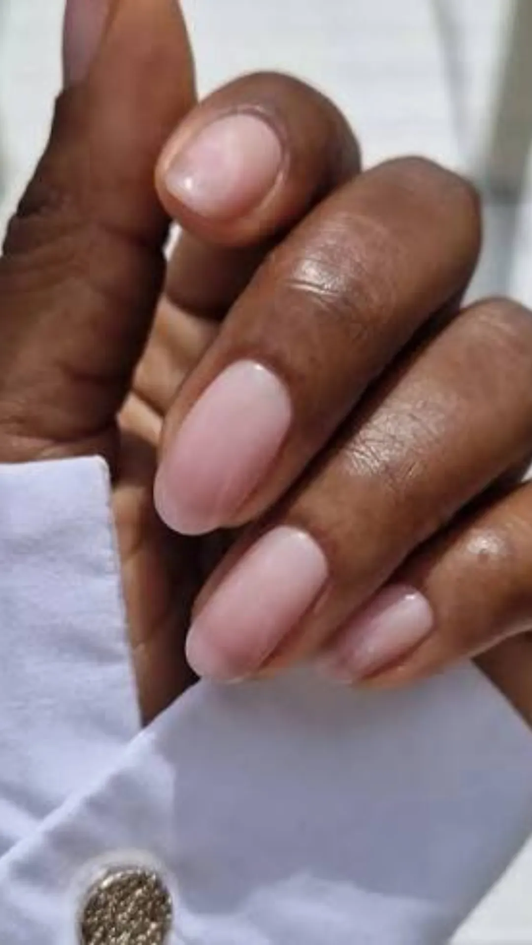 A close-up shot focusing on a neat, minimalist manicure on short, rounded natural nails. The nails are coated in a sheer, high-gloss pale pink polish that gives a clean, healthy, and ultra-natural "your nails but better" appearance. The hand is posed against the crisp white cuff of a button-down shirt sleeve featuring a textured metallic gold button.