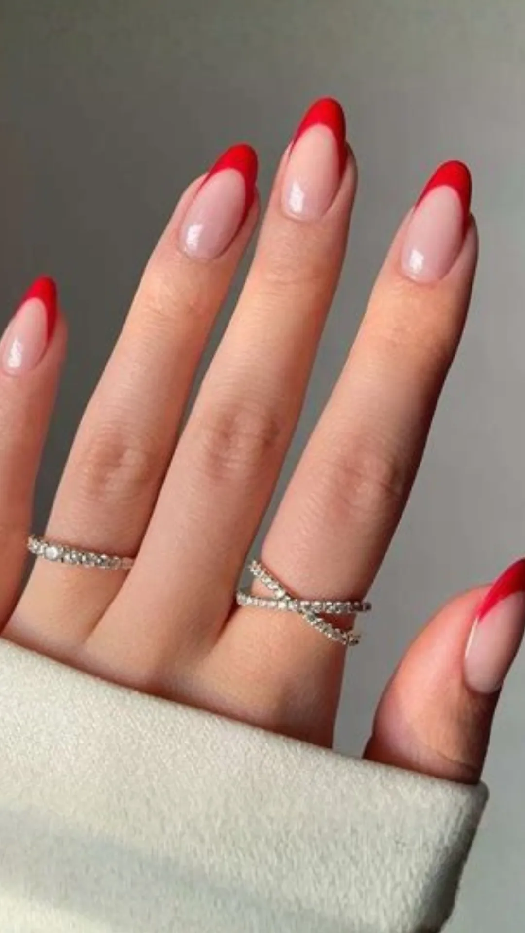 A close-up view of a hand with elegant almond-shaped nails displaying a modern twist on a classic style. The manicure features a clean, high-gloss natural nude or translucent base coat finished with thin, perfectly curved tips in a vibrant, bold cherry red polish. The model wears two delicate, diamond-encrusted silver accent rings.A close-up view of a hand with elegant almond-shaped nails displaying a modern twist on a classic style. The manicure features a clean, high-gloss natural nude or translucent base coat finished with thin, perfectly curved tips in a vibrant, bold cherry red polish. The model wears two delicate, diamond-encrusted silver accent rings.