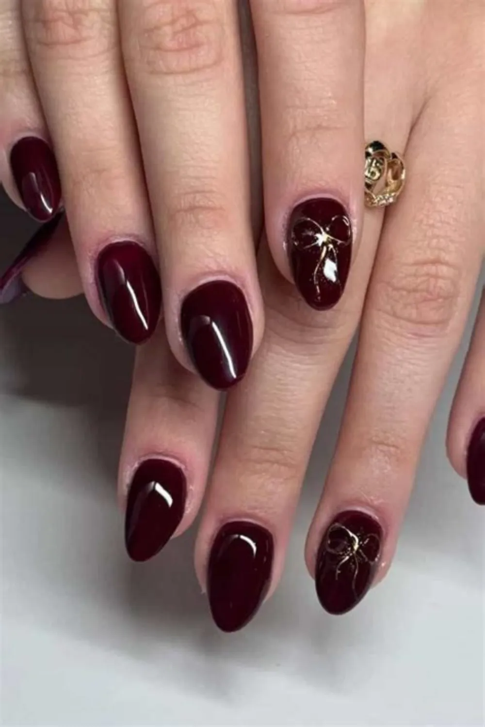 A close-up view of a chic, dark manicure on elegant almond-shaped nails. The fingernails are coated in a rich, glossy burgundy-red polish that borders on a deep wine hue. The accent nails on the ring and middle fingers are decorated near the tips with a delicate, reflective gold foil ribbon bow design that adds a subtle touch of luxury.
