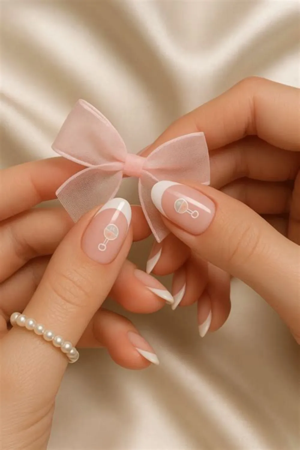 A close-up view of two hands holding a sheer pink ribbon bow against a soft cream satin backdrop. The almond-shaped nails feature an elegant, classic white French tip over a natural nude base. Decorating the thumb and index accent nails are dainty, pastel-colored baby rattle illustrations that add a subtle, sophisticated touch to the baby shower look.