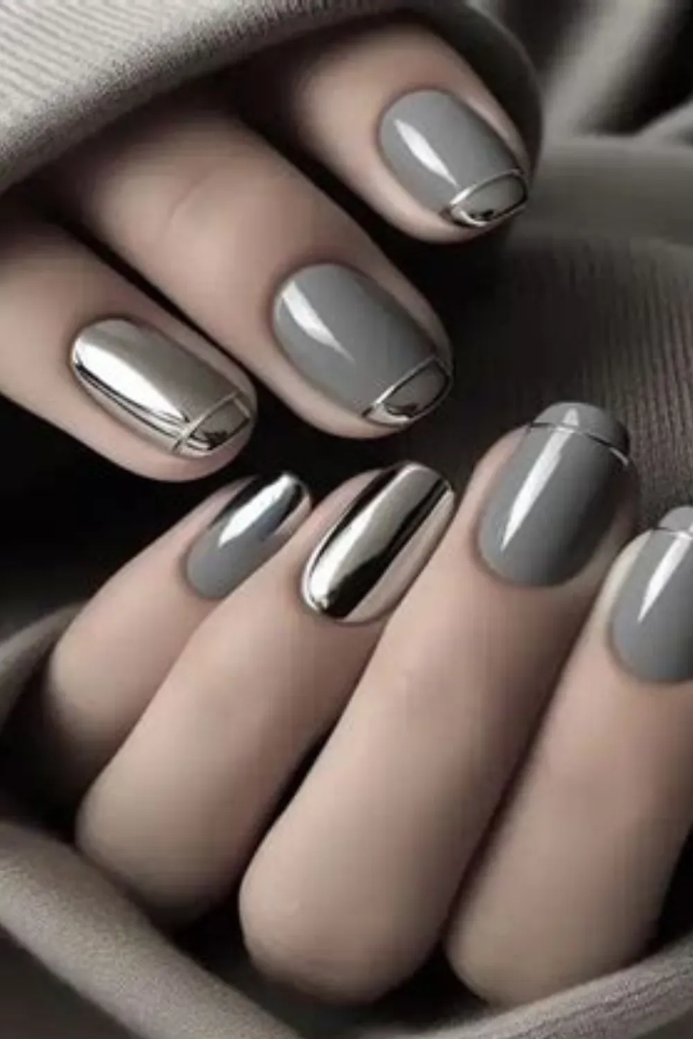 A close-up image of a hand resting inside the cozy cuff of a taupe-grey sweater, featuring a modern metallic accent manicure on short square-oval nails. The nails are painted in a glossy cool-grey polish, with select fingernails featuring liquid silver chrome accents painted in full coverage or as sharp metallic cuffs along the cuticle line.