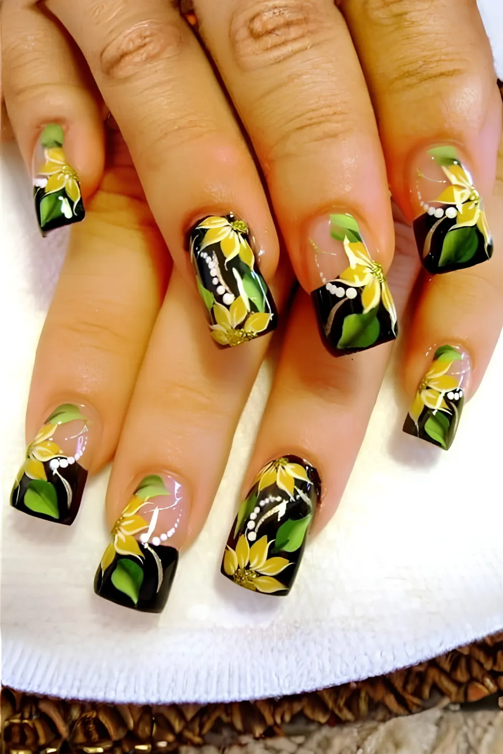 A top-down view of two hands showing square-shaped acrylic nails with an elaborate floral print design. Set against a combination of sheer and solid black tips, the nails are decorated with hand-painted tropical yellow and white Hibiscus flowers, green leaves, and delicate arched lines of white polka dots for an authentic Y2K summer vibe.