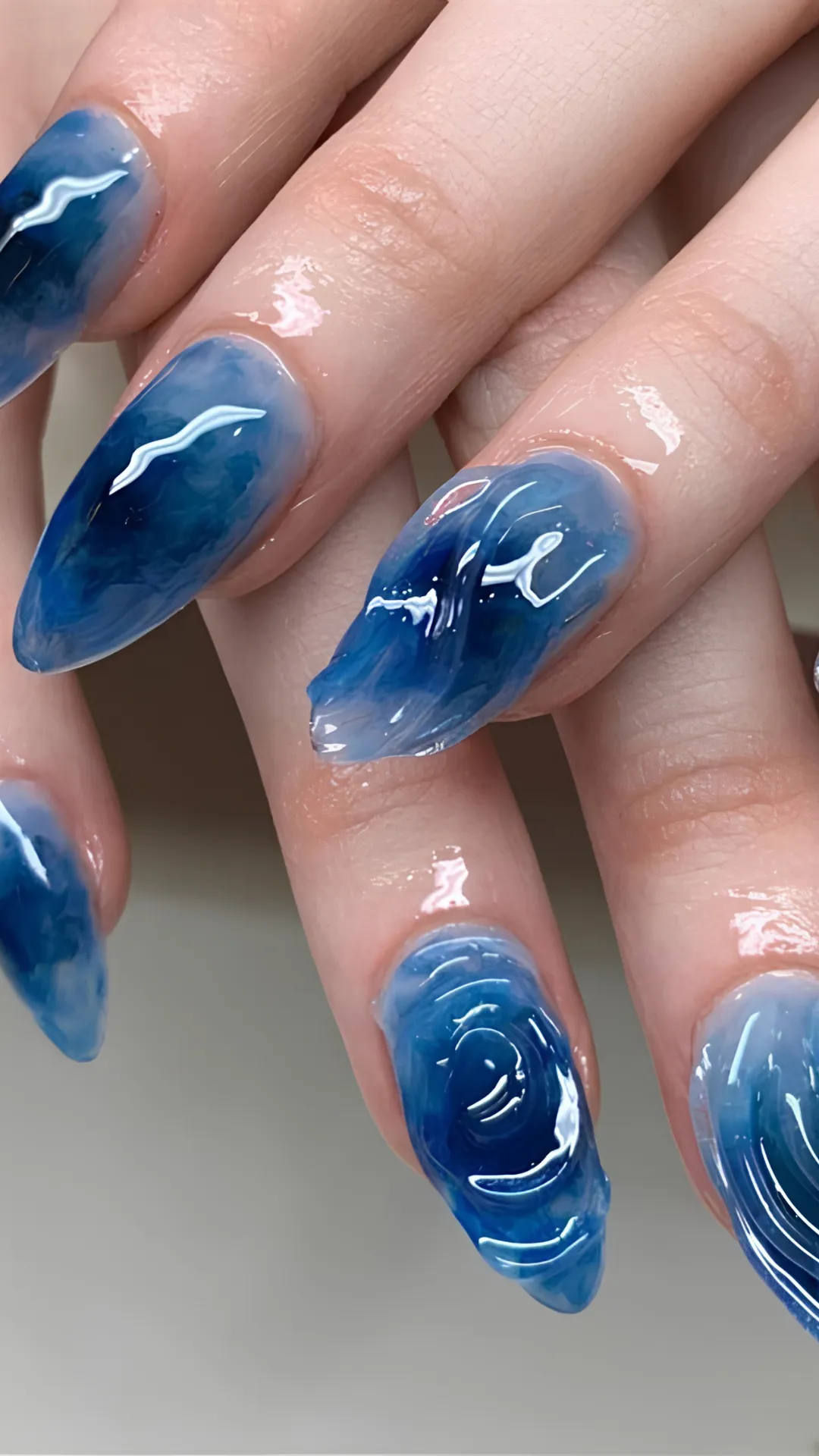 An extreme close-up of long, stiletto-shaped nails featuring a mesmerizing 3D water ripple or ocean wave effect. The nail art uses deep shades of navy blue, cerulean, and translucent white polish swirled together to mimic moving water. A thick, clear sculpted gel layer forms raised, realistic ripples and concentric circles on the nail surface.