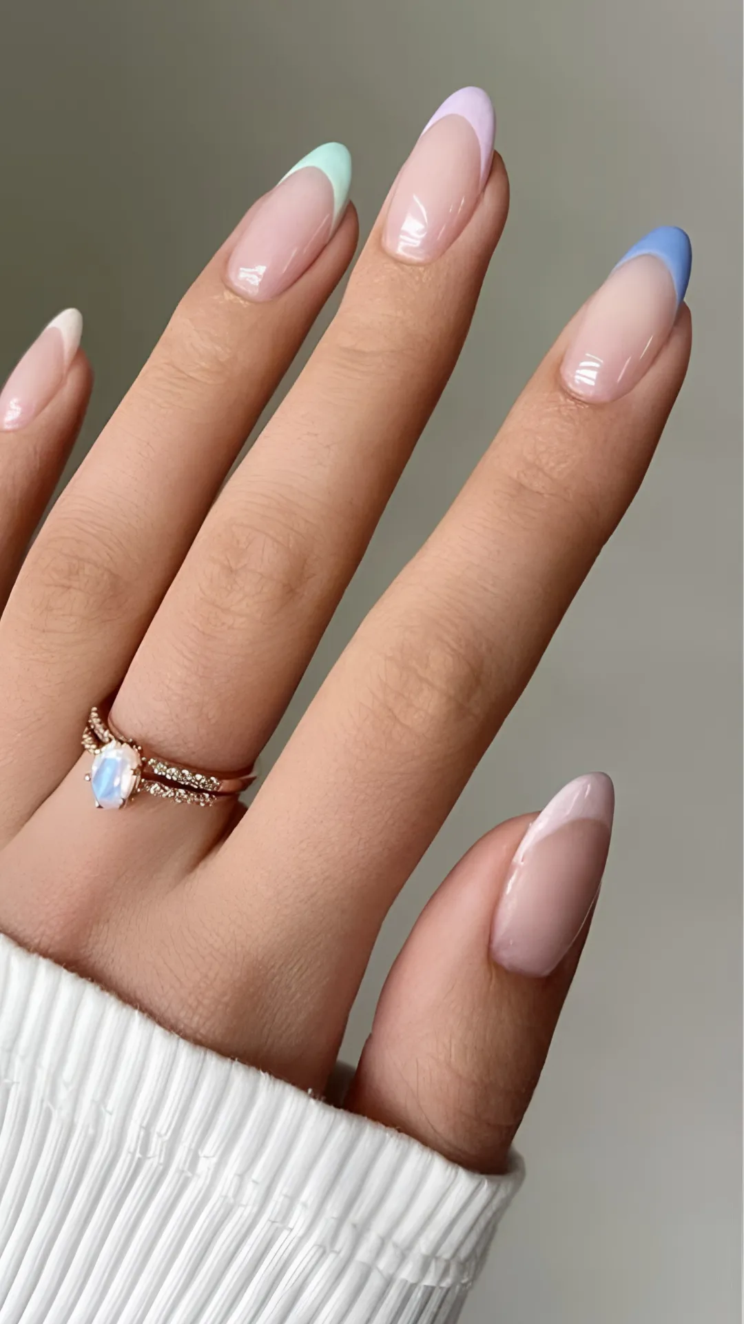 An elegant, minimalist spring manicure showcased on long, oval-shaped nails. The design is a modern twist on the classic French manicure, featuring a sheer, high-gloss nude base coat with ultra-thin pastel-colored tips. Each fingernail sports a different hue, including mint green, lilac, cornflower blue, and soft baby pink, complemented by a dainty gold gemstone ring worn on the ring finger.