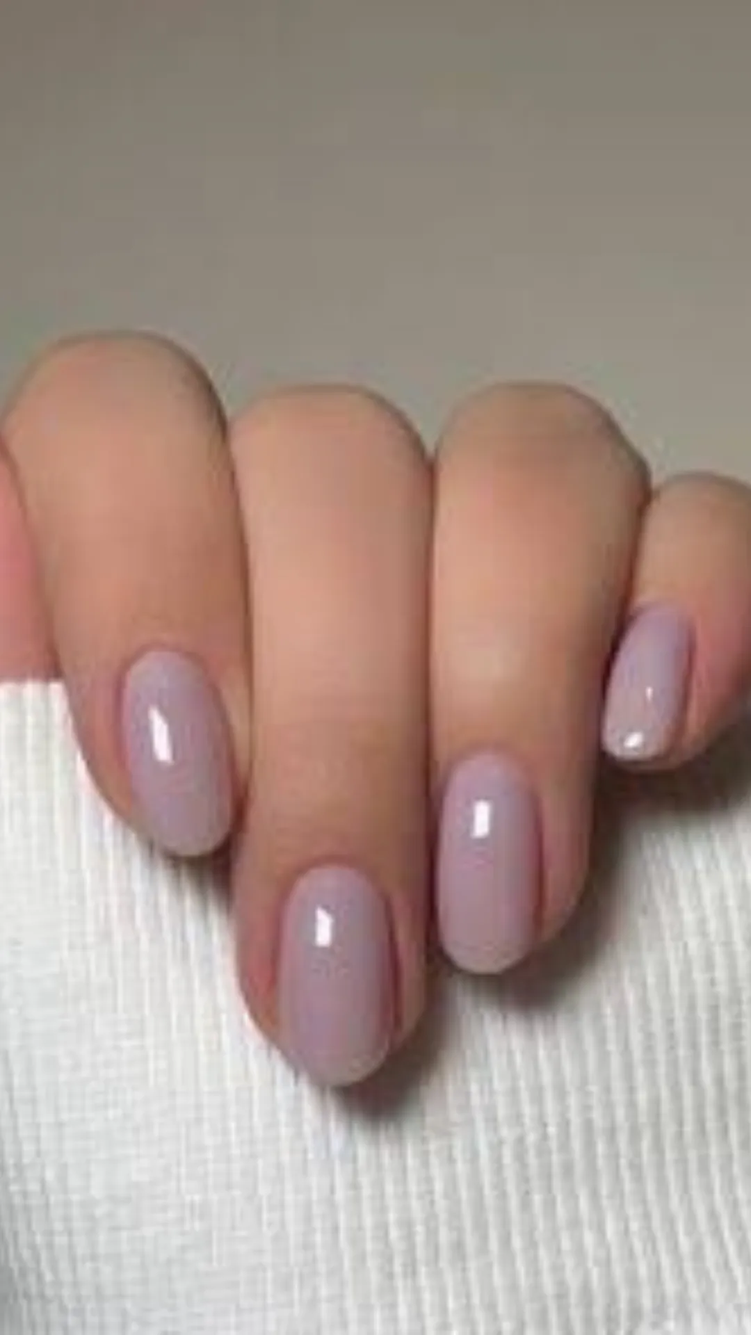 A detailed view of a classic, understated manicure on short, oval-shaped nails. The fingernails are painted in a smooth, high-gloss lavender-grey or cool taupe polish that offers a sleek, monochromatic look suitable for professional wear. The hand is set against the clean lines of a white ribbed fabric background, emphasizing a polished and put-together style.