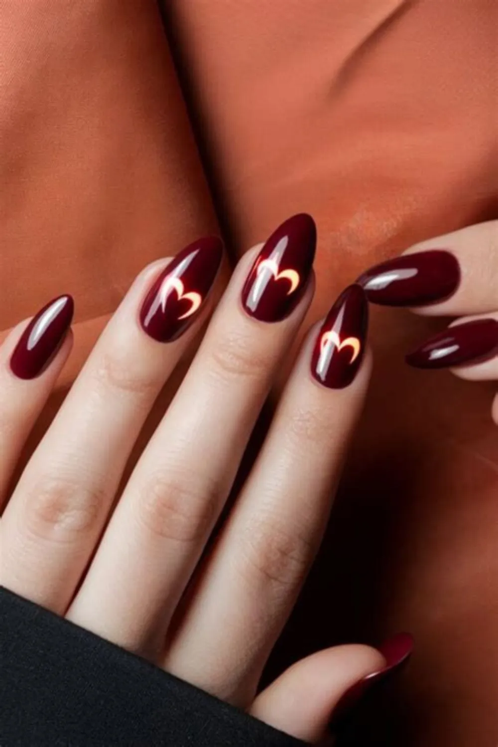 A detailed shot of a hand showcasing a bold negative space heart design on long, oval fingernails. The background polish is a high-gloss, deep maroon or dark cherry red lacquer. The center of the index, middle, and ring fingers features a precise cutout silhouette forming an outlined, glowing heart shape that reveals a bright coral-red tone underneath.