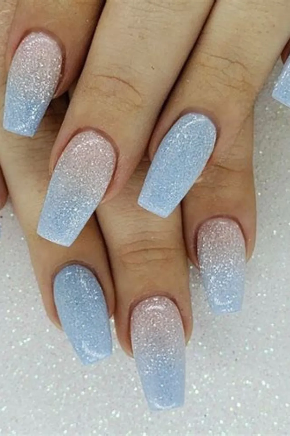 A close-up view of long, coffin-shaped nails featuring a dazzling glitter ombré baby boy design. The nail art seamlessly transitions from a sheer nude base at the cuticles into a soft pastel blue at the tips, heavily blended with fine, shimmering silver micro-glitter throughout to give a magical, frosted appearance against a glittering background.