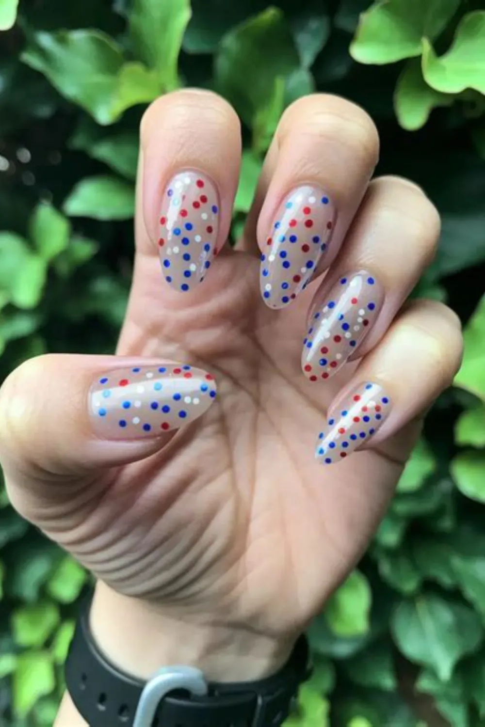 A close-up photograph of a hand held against a lush green leafy background, displaying a playful confetti-style 4th of July manicure. The long, almond-shaped nails feature a sheer, high-gloss natural nude base covered from cuticle to tip in tiny, randomly scattered polka dots painted in classic red, white, and royal blue.