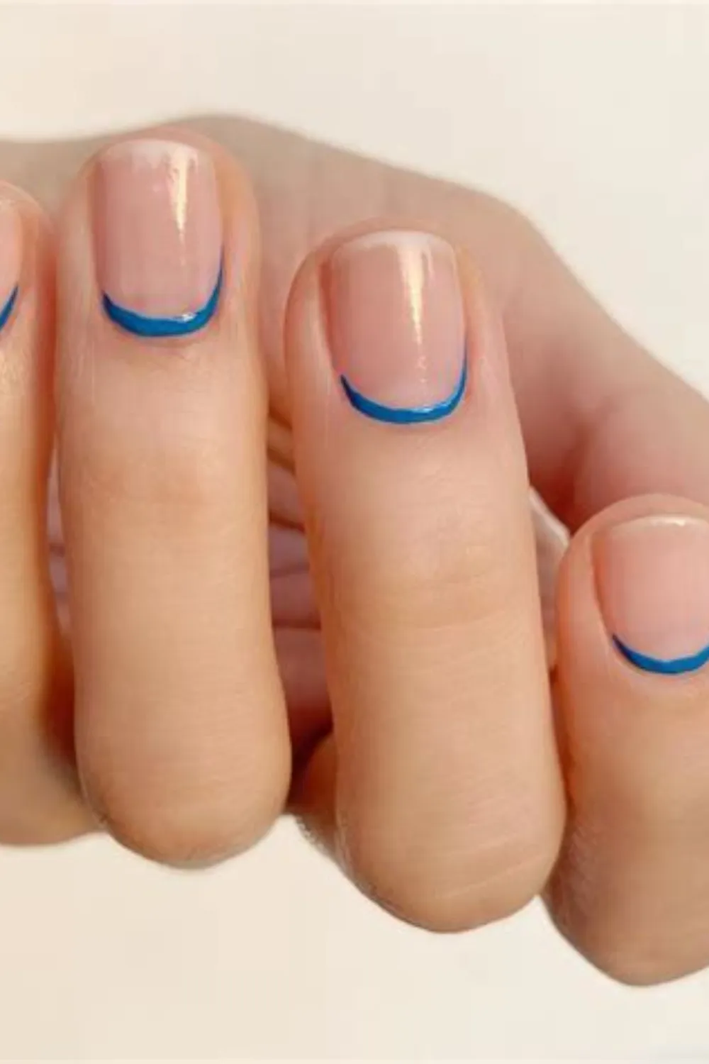 A detailed macro view of short, rounded natural nails displaying an unexpected twist on the French manicure. The nails are covered in a glossy, translucent nude base polish, accented by thin, vibrant bright electric-blue lines painted precisely along the curved cuticle lines (reverse French tips) instead of the top edges.