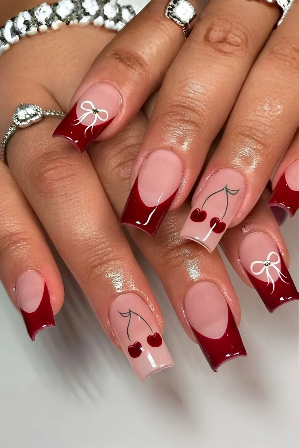 A detailed photograph of a hand wearing a heart-shaped diamond ring, displaying a retro coquette-meets-Y2K French manicure on square coffin nails. The nails feature deep cherry-red French tips; the ring and middle fingernails are decorated with fine white ribbon bow outlines, while the accent nails feature hand-painted red cherries on green stems over a nude base.