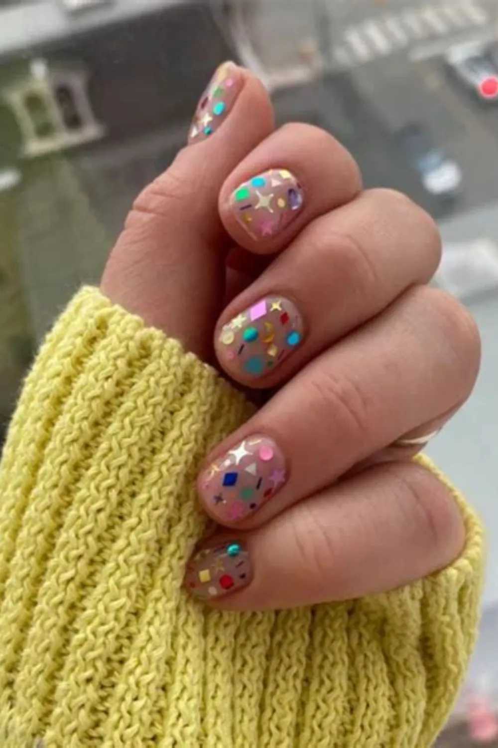 A close-up photograph of a hand emerging from the sleeve of a pastel yellow cable-knit sweater, displaying a confetti-inspired short manicure. The neat, rounded nails are painted with a clear, high-gloss base coat and covered in a whimsical array of colorful metallic confetti sequins shaped like stars, diamonds, moons, and circles
