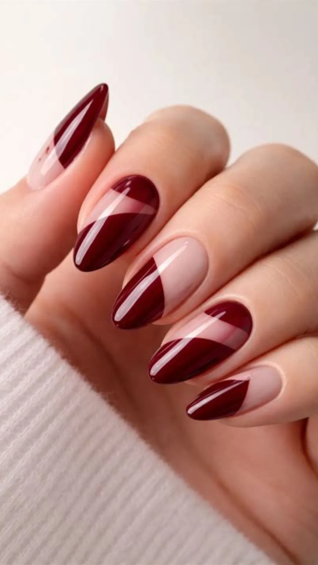 Geometric negative-space nail art on long almond nails, showcasing clean diagonal blocks of glossy burgundy polish paired with bare, natural nail sections.