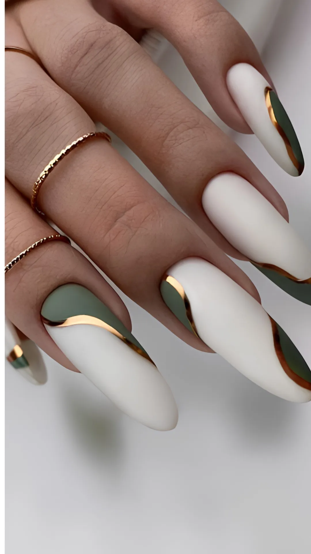 Artistic matte white stiletto nails decorated with elegant, flowing abstract swirls of olive green and outlined with bold metallic gold chrome linework.
