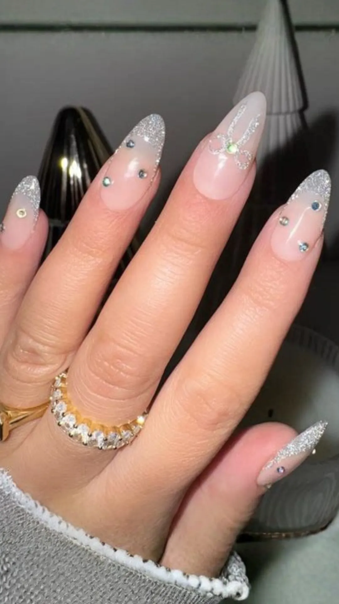 Romantic coquette nail art showcasing long almond nails with a glossy nude base, delicate silver glitter French tips, scattered rhinestones, and a hand-painted silver glitter bow.