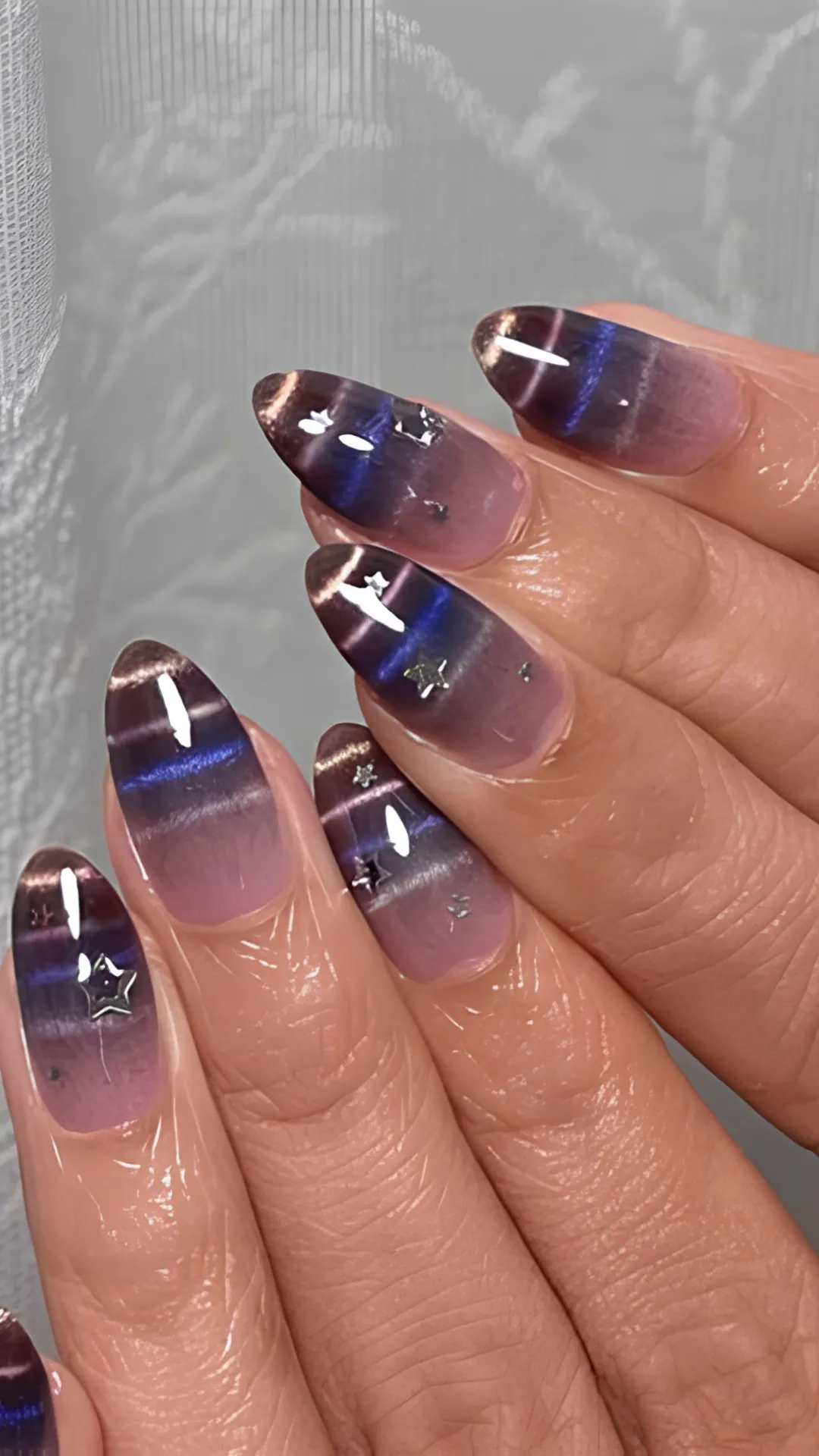 Close-up view of a hand displaying an atmospheric Korean galaxy-inspired cat-eye manicure. The long, almond-shaped nails feature a dark purple and deep indigo magnetic shimmer stripe running horizontally across a translucent mauve base. Tiny, reflective silver star studs and miniature metallic flecks are embedded into the polish to create a starry night sky effect.