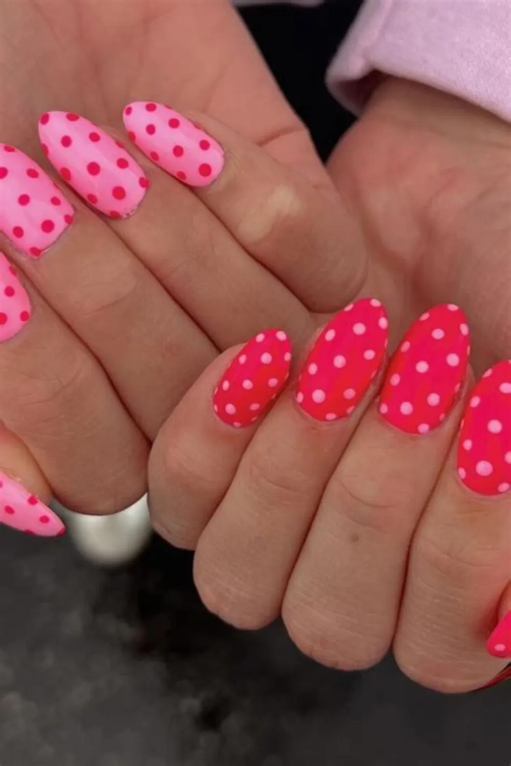 A vibrant close-up photograph displaying a playful, retro-inspired polka dot manicure. The almond-shaped nails alternate between a bright bubblegum pink base covered in bold, evenly spaced hot red polka dots, and a hot red base covered in contrasting pastel pink polka dots, creating a fun, energetic, and high-visibility color palette.