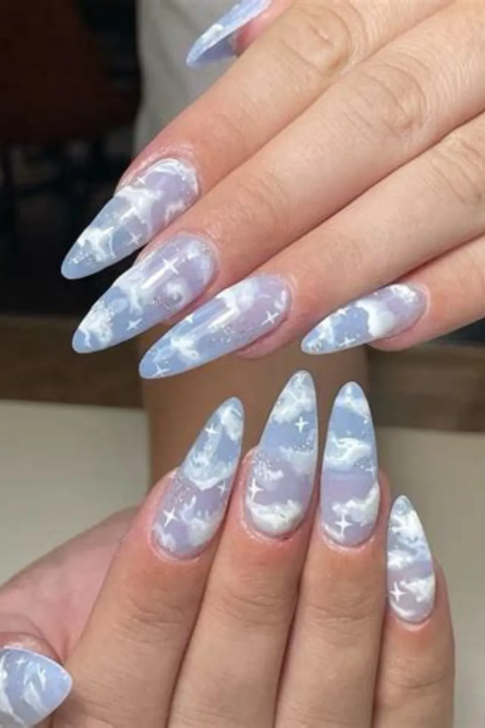 A detailed photograph of two hands featuring a dreamy, celestial cloud manicure on long, stiletto-shaped nails. The design uses a semi-translucent, milky lavender and sky-blue base coat layered with soft, wispy white clouds, fine silver shimmer, and hand-painted white four-point starbursts spread across every nail for an ethereal aesthetic.