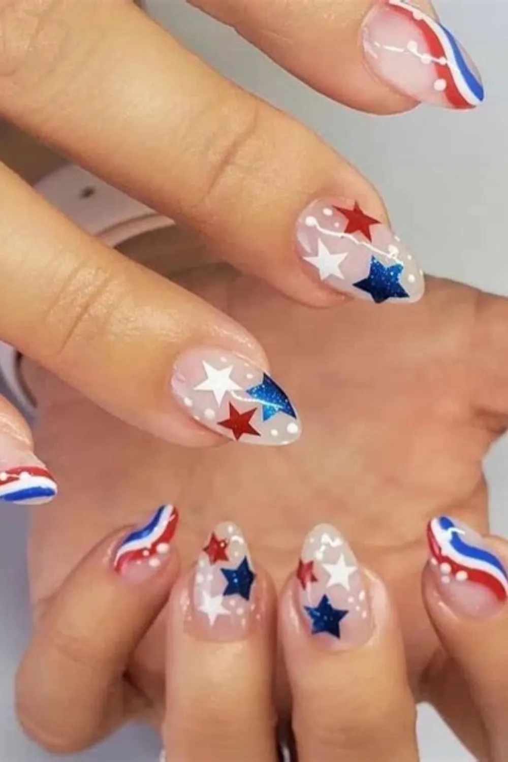 A close-up shot of two hands displaying an elegant patriotic nail art design on almond-shaped nails. The manicure uses a semi-translucent nude base; accent nails feature crisp red, sparkling blue, and bright white stars surrounded by fine white specks, while the surrounding nails are framed with flowing red, white, and blue wave lines.