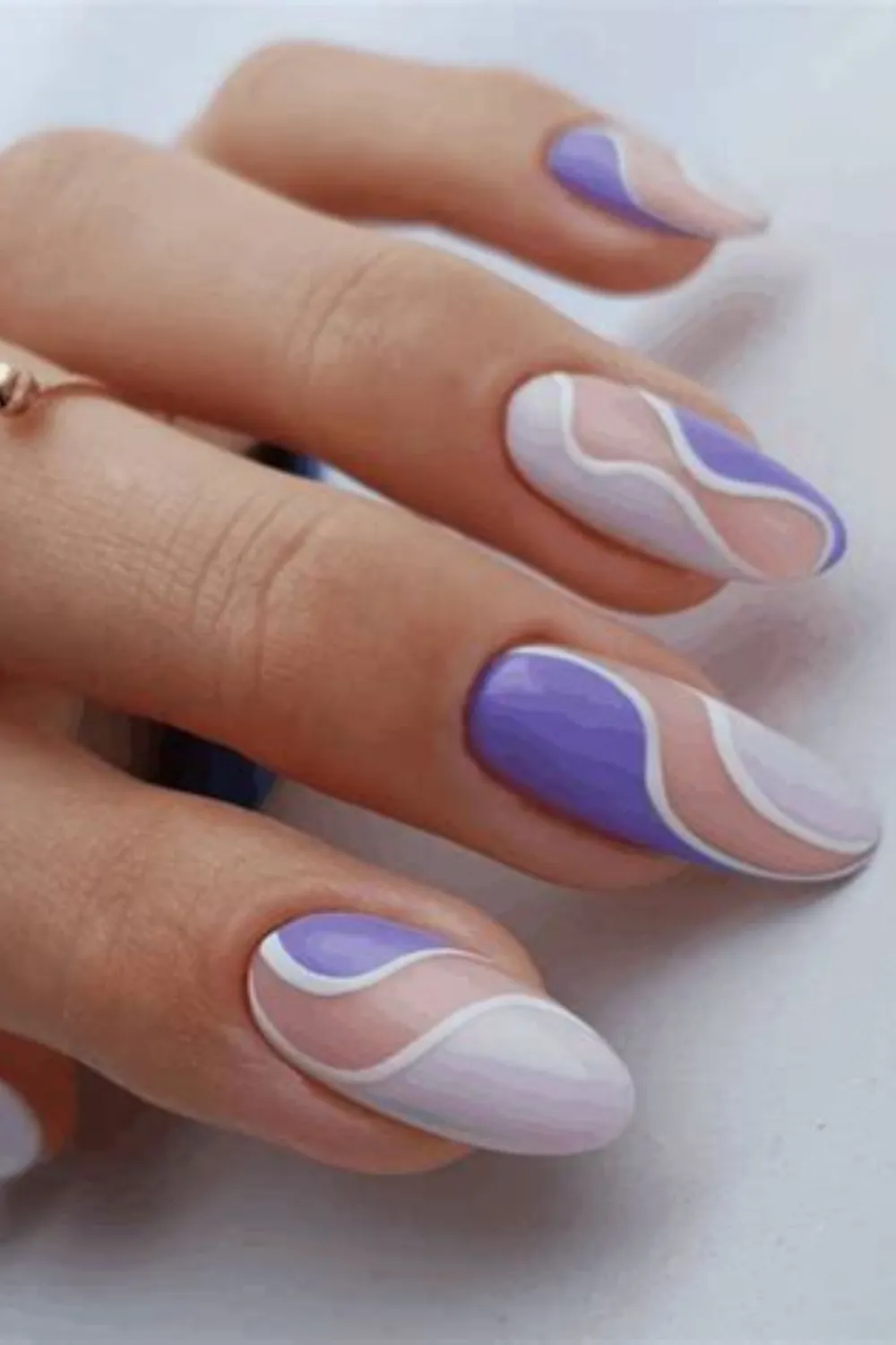 A close-up photograph showcasing long, oval-shaped nails with an artistic retro-swirl pattern. Over a sheer nude base, smooth, flowing abstract wave lines in pastel lavender, periwinkle blue, and stark white wind across each nail bed, creating a seamless, organic graphic design with clean negative space.