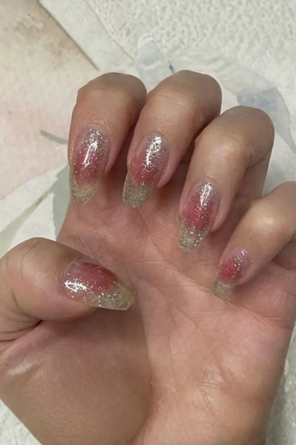 A close-up shot of a hand displaying transparent jelly extension nails with a sparkling pink aura effect. The long, tapered nails feature a clear builder gel base with a airbrushed blush of shimmering pink glitter concentrated near the center of each nail bed, giving a translucent glass-like appearance filled with silver micro-glitter.