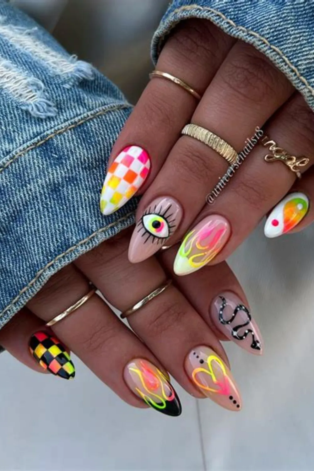 A detailed view of two hands wearing a ripped denim jacket, displaying a bold, neon festival-inspired manicure on almond-shaped nails. Over a nude base, each finger features vibrant graphic art, including neon flames, a psychedelic evil eye with long lashes, a black-and-pink checkerboard pattern, a coiled black snake, and a glowing neon heart outline.
