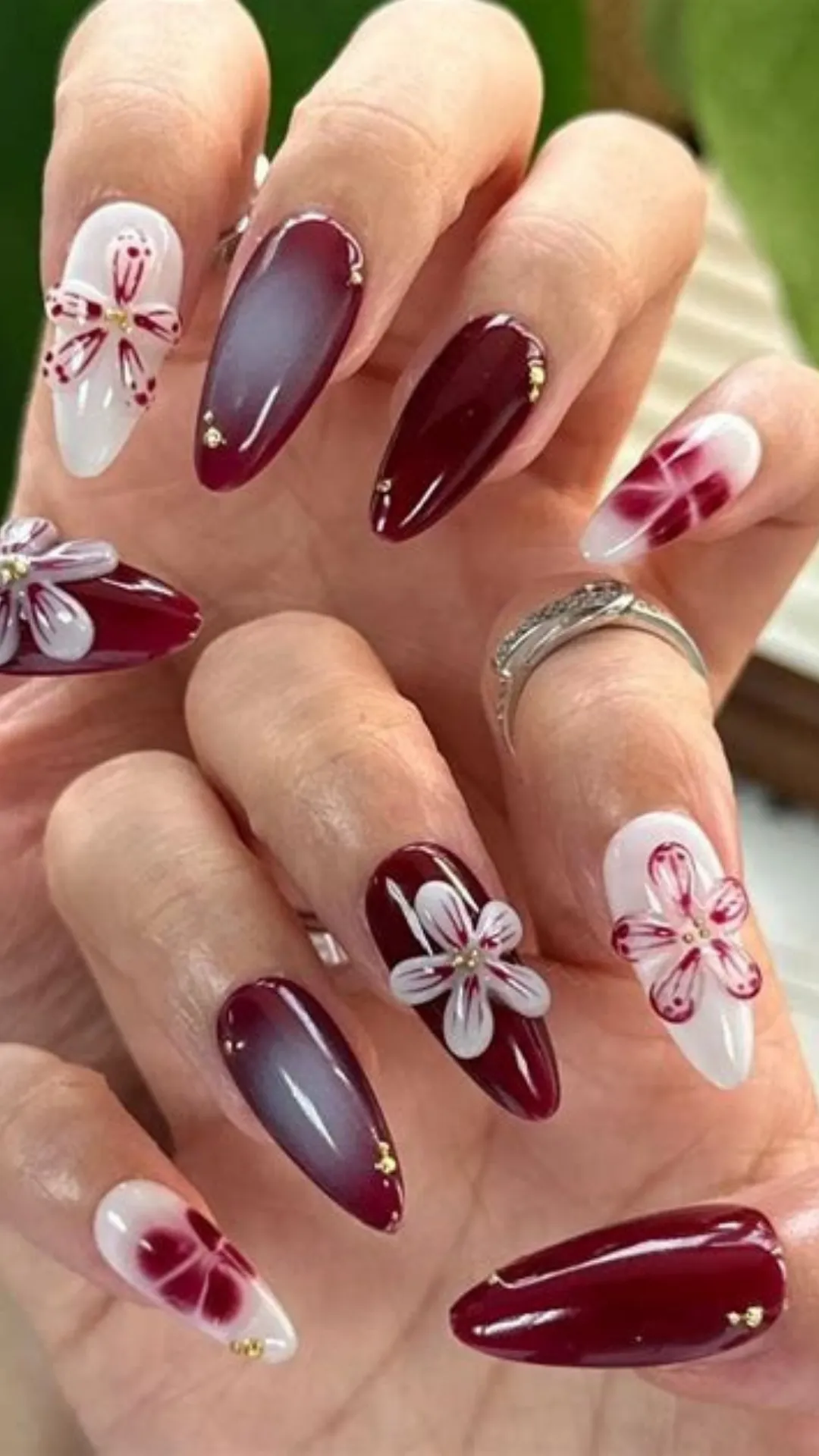 Exquisite 3D floral nail design featuring glossy burgundy nails with tiny gold studs, combined with white accent nails showcasing raised, sculpted red and white flower petals.