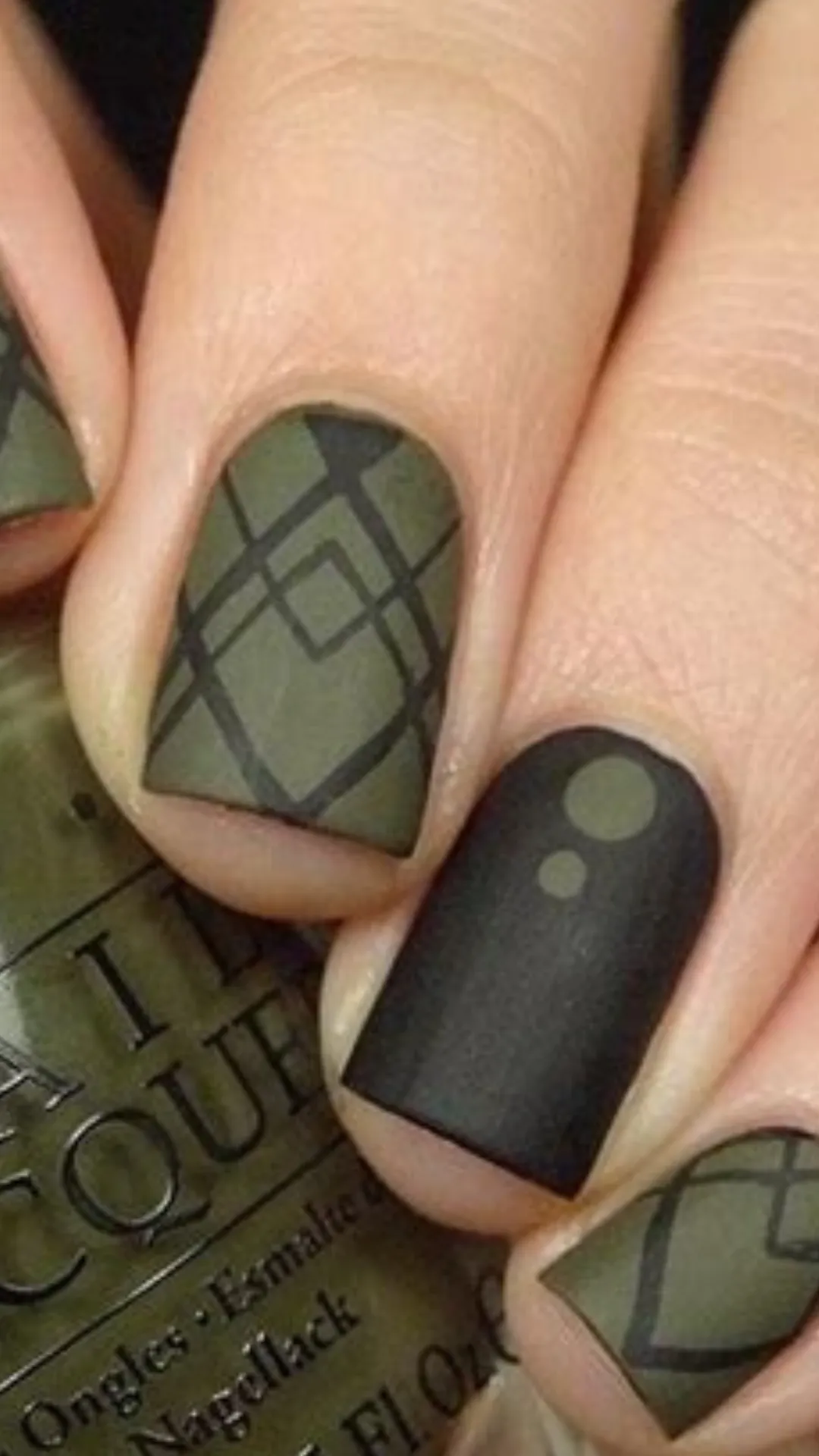 A close-up of short square nails featuring a matte olive green base painted with a dark green plaid pattern, paired with a solid matte black accent nail detailed with green dots.