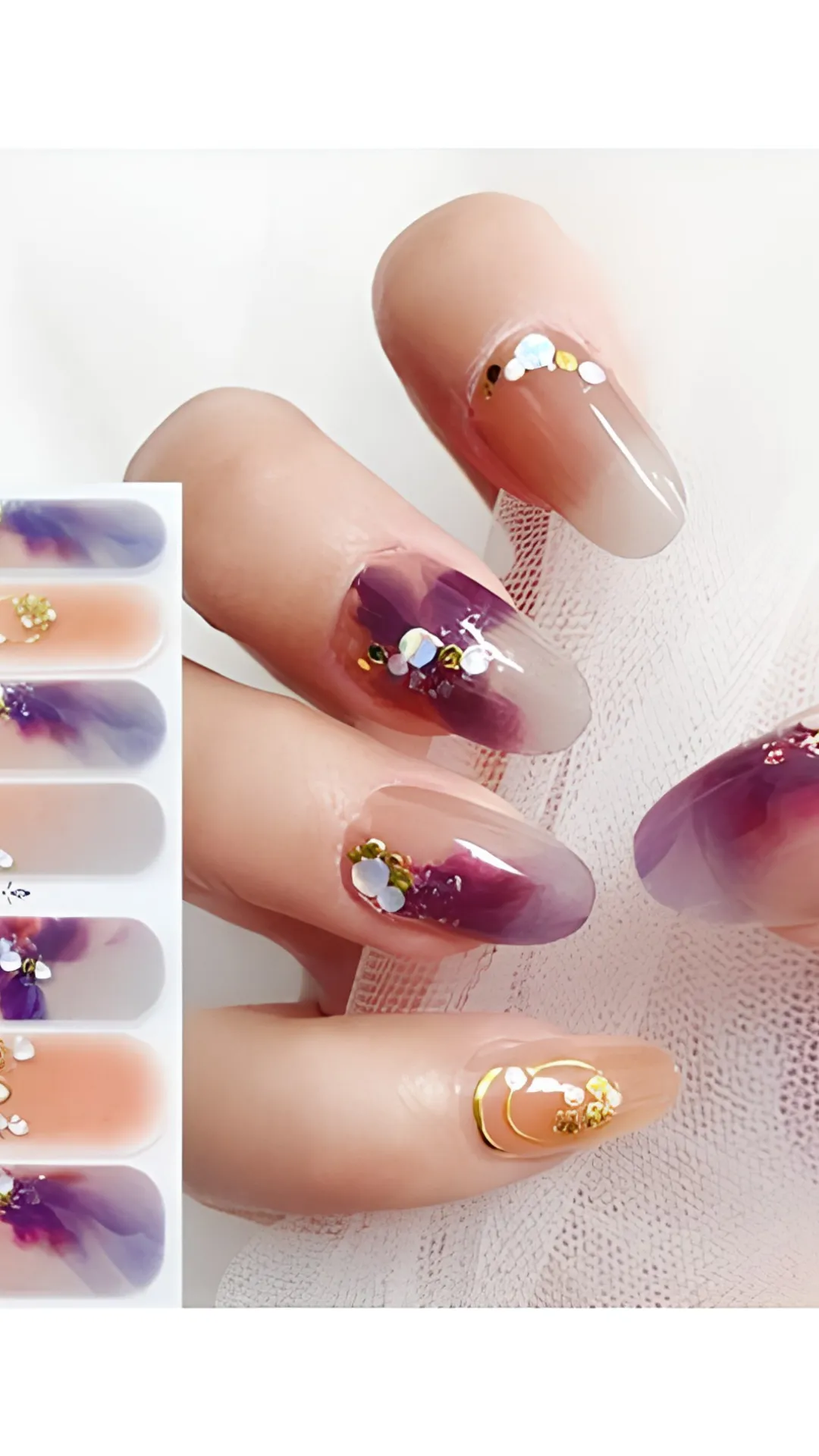 A top-down view of a hand showcasing a delicate floral Korean nail wrap design alongside its packaging strips. The long oval nails feature a sheer nude base decorated with watercolor-style deep purple flower petals, gold metallic foil wire circles, tiny white floral accents, and chunky iridescent glitter flakes that catch the light beautifully.