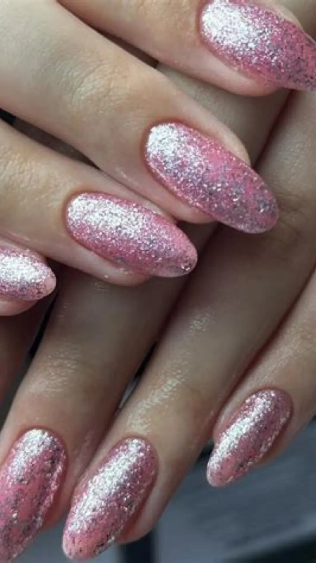 This close-up shot focuses on hands showcasing high-impact sparkle on elegant short almond-shaped nails. The nails are completely covered in dense, reflective pink and silver holographic glitter polish, creating a dazzling effect under the light. The glitter is evenly distributed, achieving a solid, jewel-like finish while maintaining the clean almond shape. The hands are subtly posed against a dark blurred background, emphasizing the reflective, glamorous sparkle. This versatile design offers a striking yet sophisticated everyday look that transitions effortlessly to evening wear.