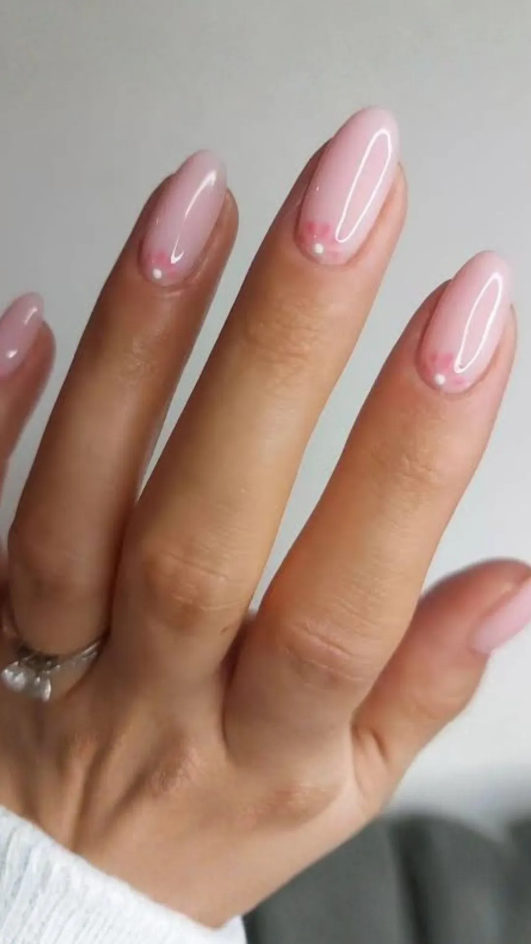 A close-up photograph of a hand displaying long, oval-shaped nails with a minimalist cherry blossom design. The nails feature a high-gloss, semi-sheer bubblegum pink base coat. Near the cuticle area of each finger, a simple three-petaled pink flower outline is painted, centered with a single, clean white dot. The person is wearing a silver engagement ring with a visible gemstone.