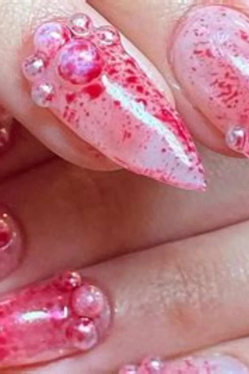 An extreme macro photograph focusing on a dramatic, avant-garde textured manicure on long, sharply pointed stiletto nails. The design features a semi-translucent base covered with a splattered, abstract texture resembling deep red watercolor veins or crimson stone marble. The cuticles are heavily embellished with clusters of matching red and clear 3D rhinestone gems.