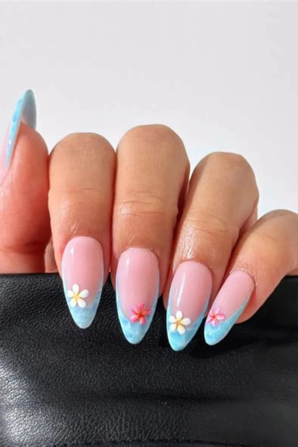 A close-up shot of a hand holding the edge of a black leather item, displaying long almond-shaped nails with an ocean-themed French manicure. The tips feature a wavy, light blue water-texture design, decorated with tiny 3D tropical plumeria flowers in white and pink centered on each nail tip over a natural glossy nude base.