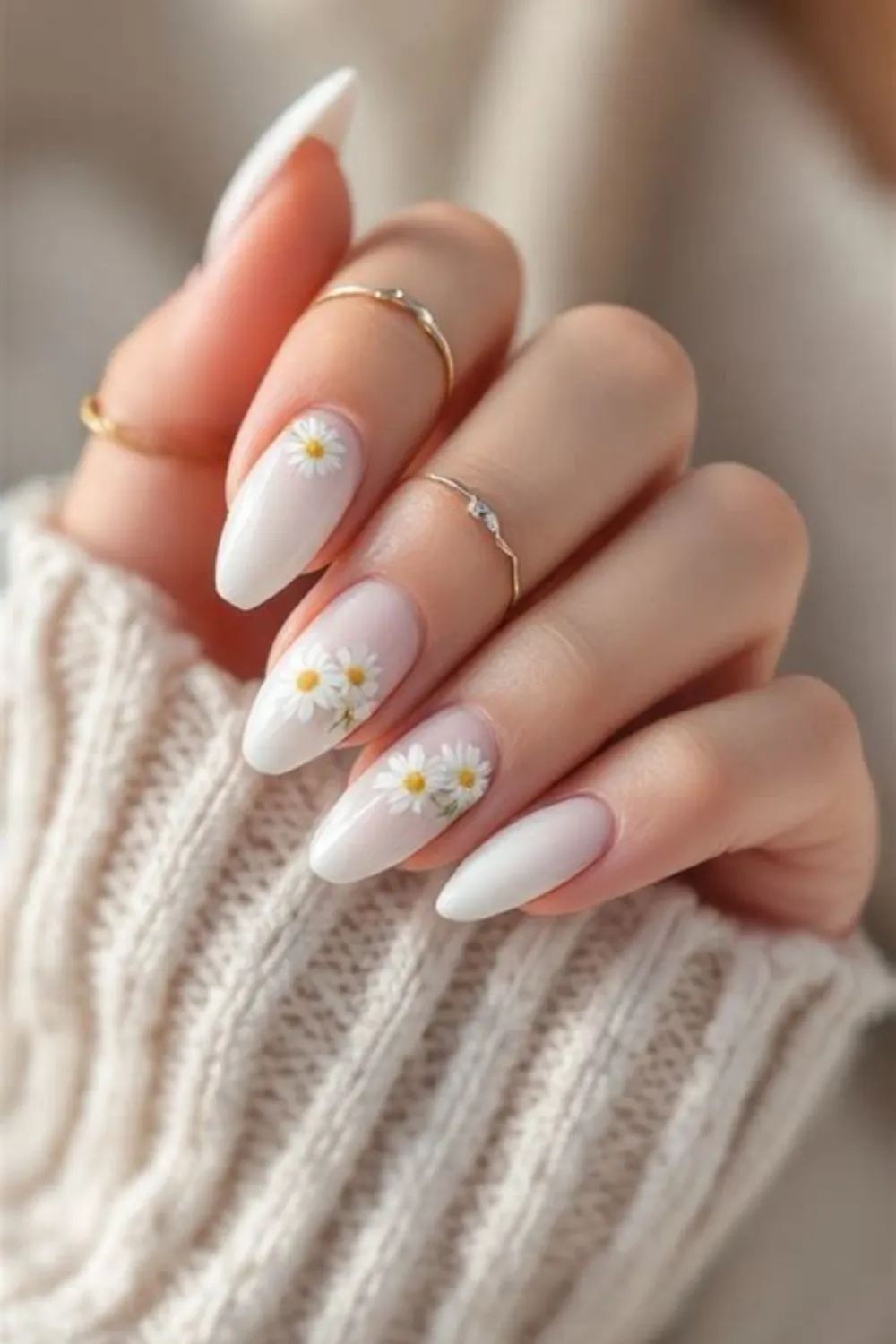 A close-up view of a hand with long, almond-shaped nails peeking out from the sleeve of a cream-colored cable-knit sweater. The nails feature a milky translucent white gel base decorated near the cuticles and centers with dainty, hand-painted white daisies with bright yellow centers, accessorized by thin gold rings.
