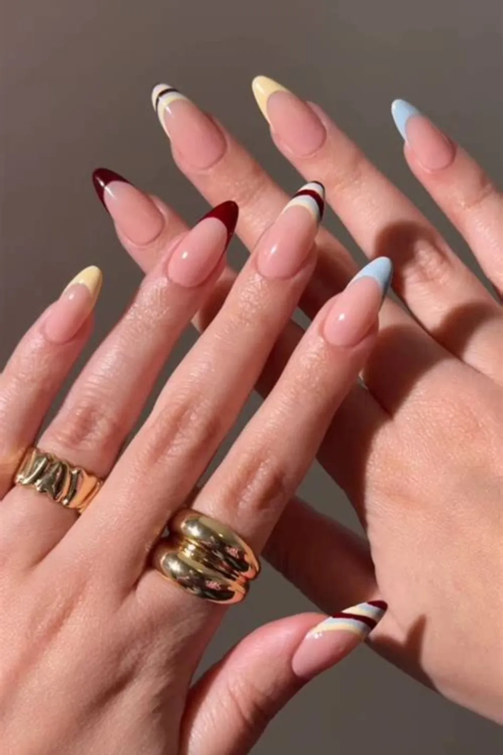 A close-up photograph of two hands crossed over each other, displaying a chic multi-colored French manicure on long, oval-shaped nails. Over a glossy natural nude base, the thin French tips feature alternating retro color combinations, including sky blue, butter yellow, deep burgundy, and tri-color stacked arcs, paired with thick, polished gold dome rings.