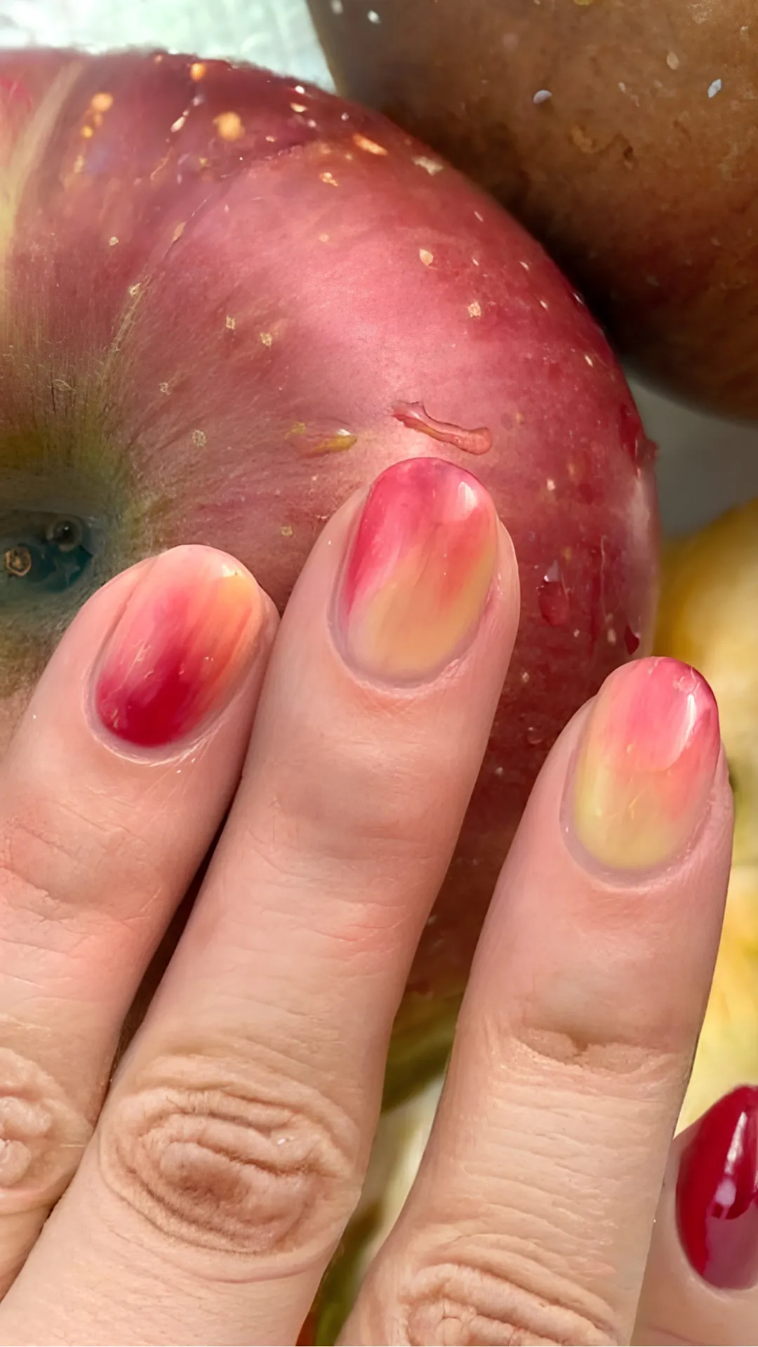 A close-up shot of a hand resting against a vibrant red apple to highlight a fruit-inspired Korean manicure. The short, rounded nails feature a glossy gradient ombré design that transitions from a pale, soft yellow at the cuticles to a rich strawberry red at the tips, perfectly mimicking the natural coloration of a ripe apple.